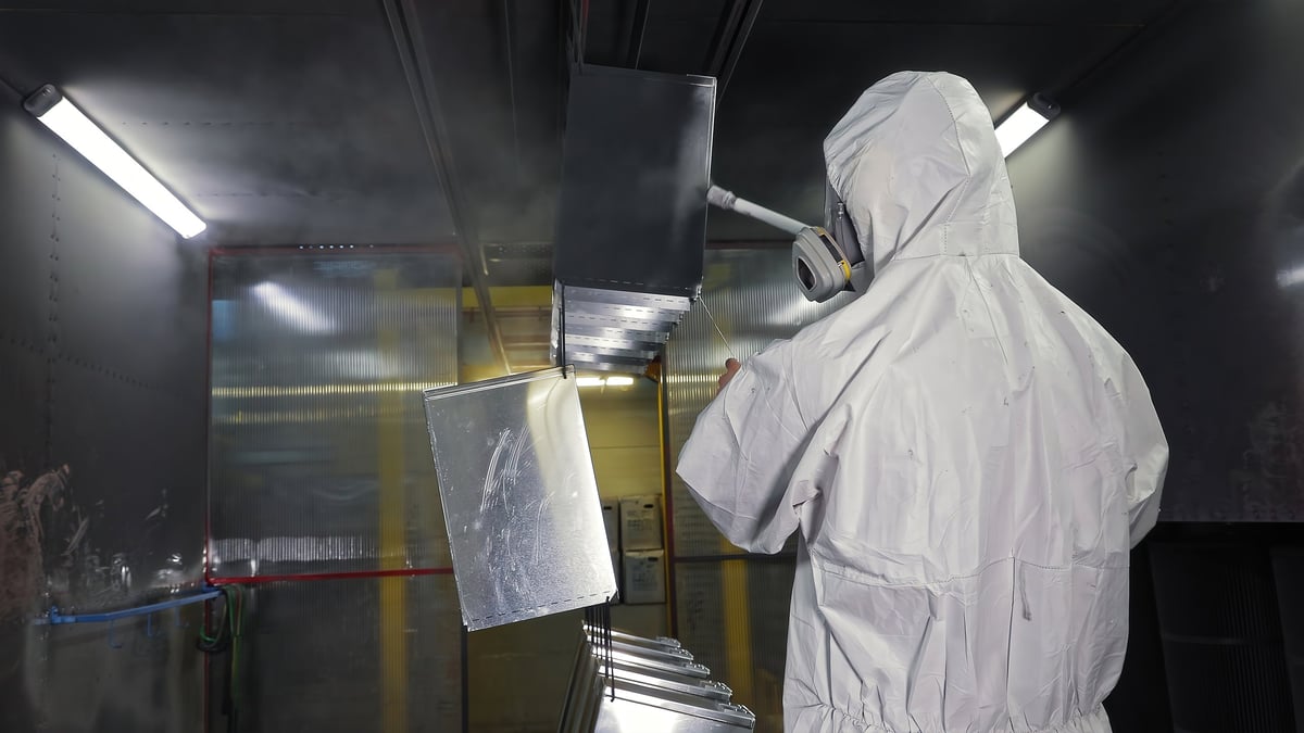 Technician in a protective suit spray coating metal parts inside a spray booth